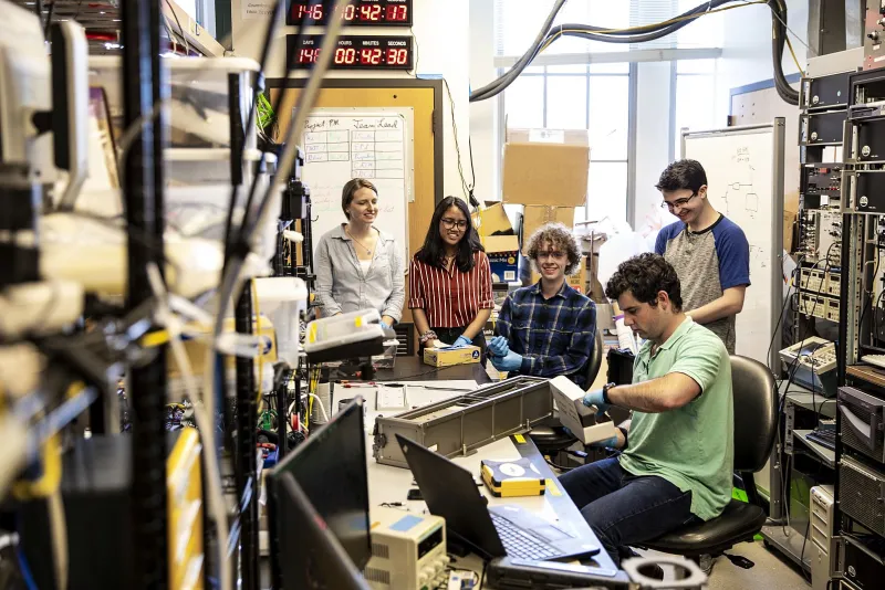 Team members Paige Northway, Anika Hidayat, John Correy and Eli Reed (back row, left to right) watch in June as Henry Martin of Nanoracks does a “fit test” to ensure that the satellite fits inside the silver box